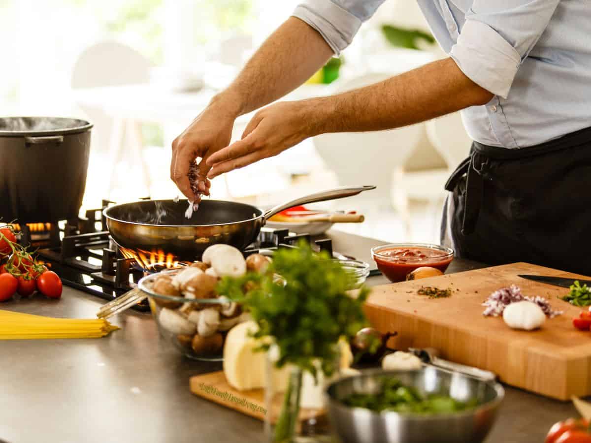 Fresh ingredients in bowls or on cutting board surround cooktop with skillet. Man's hands placing some of the prepared vegetables into the skillet.