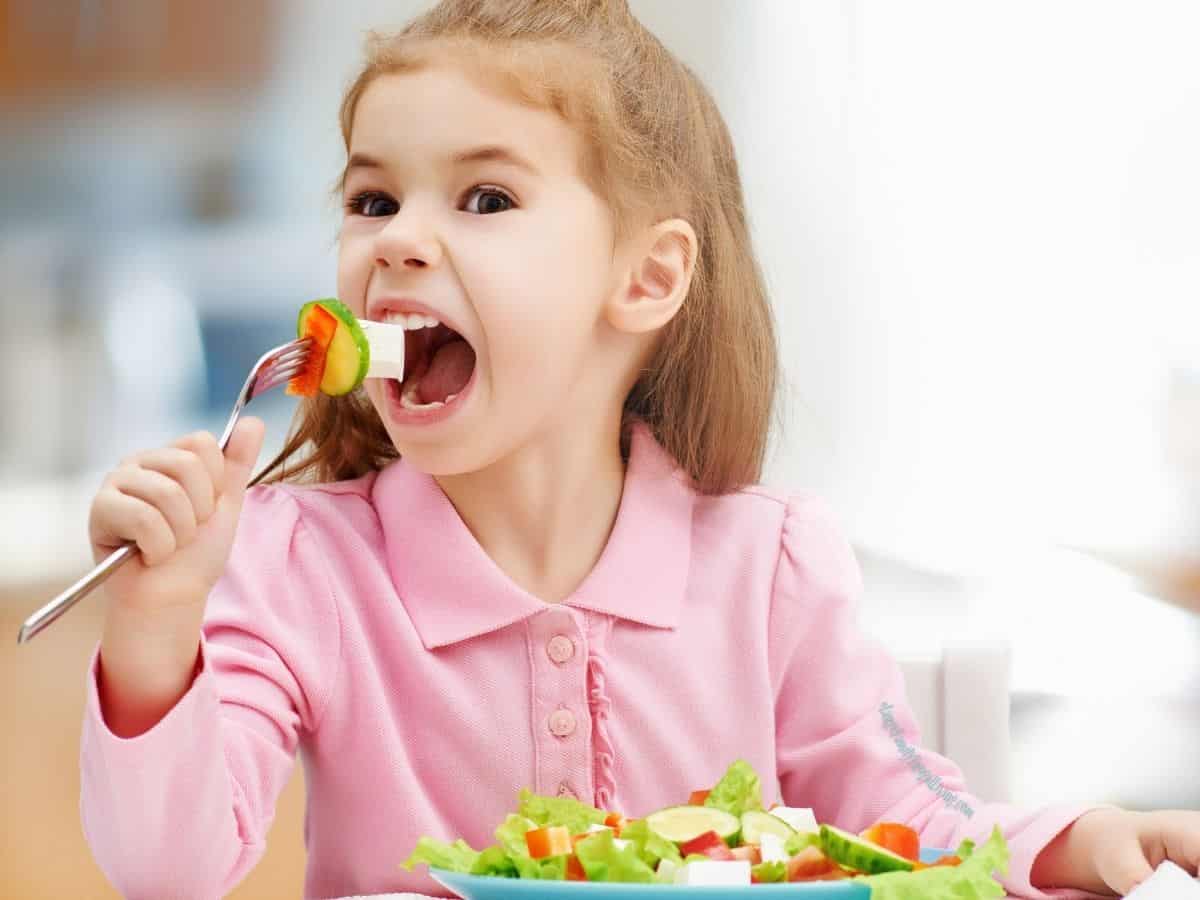 Little girl in pink top sitting at a table with a plate - putting a forkful of veggies in her mouth