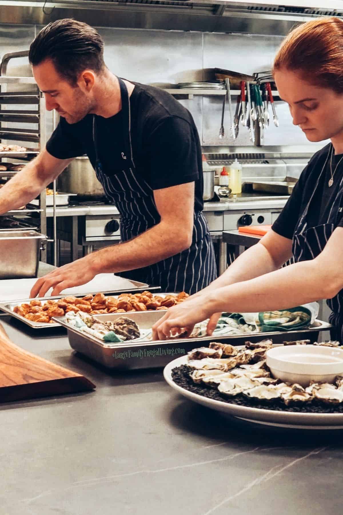 Man and woman wearing black striped aprons arranging foods on wood board