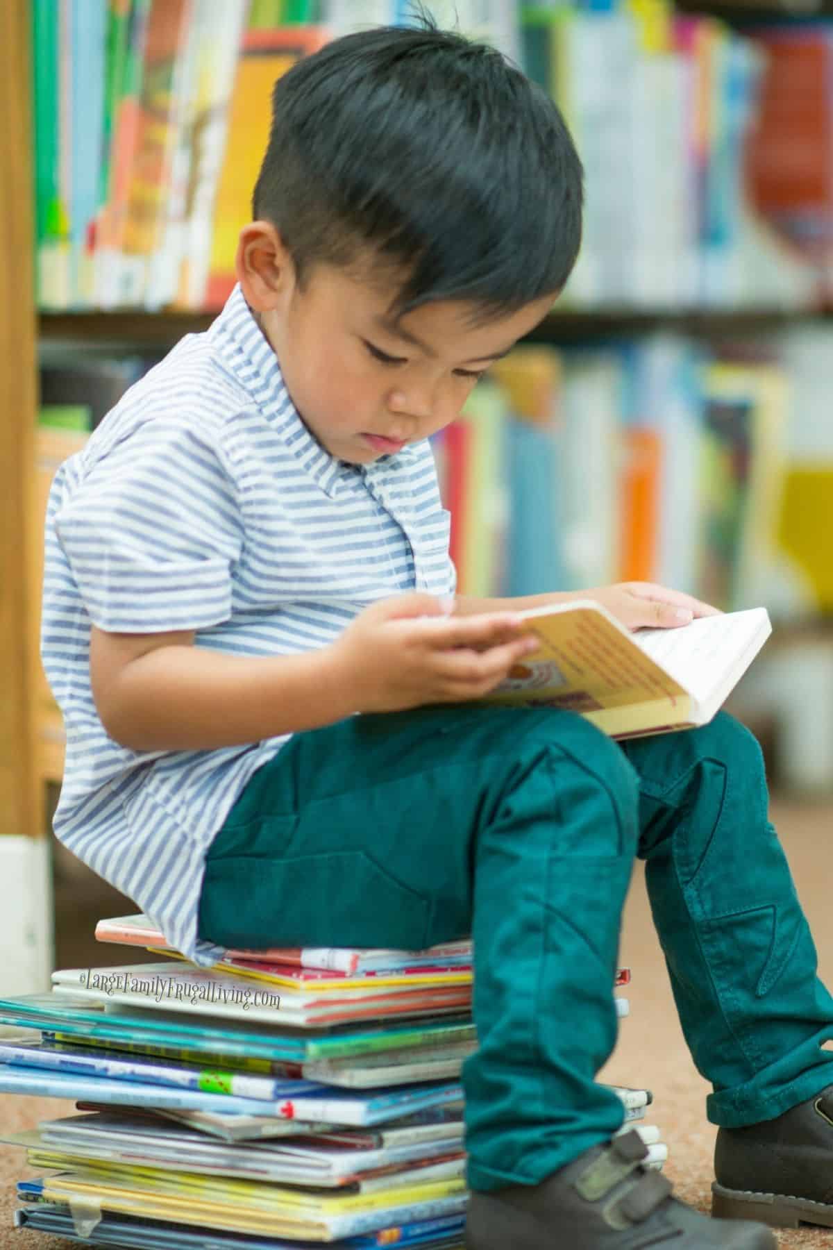 Young boy sitting on stack of books while reading a book