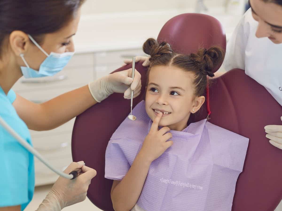 Smiling girl in treatment chair at dentist office pointing at her missing tooth with dentist and assistant