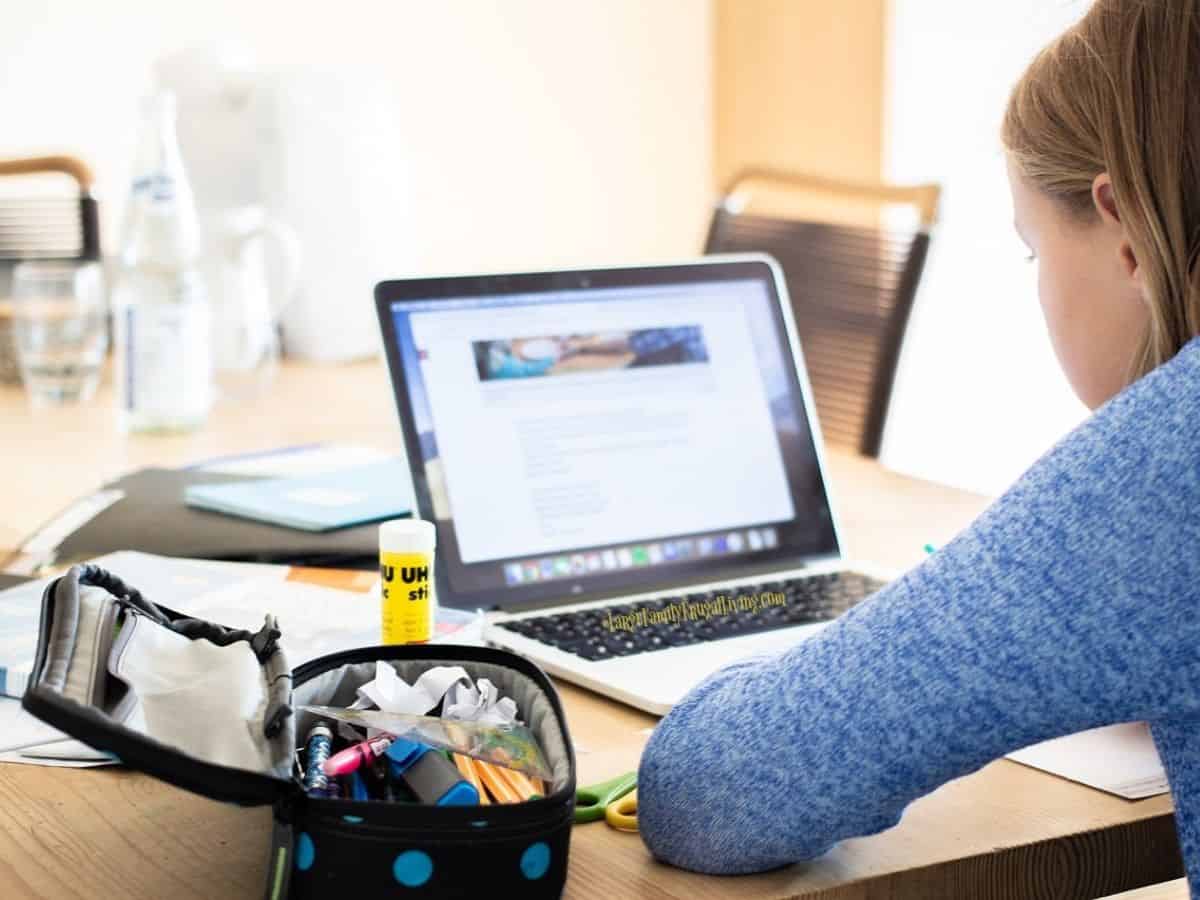 Girl working on laptop sitting at a kitchen table with school supplies beside her